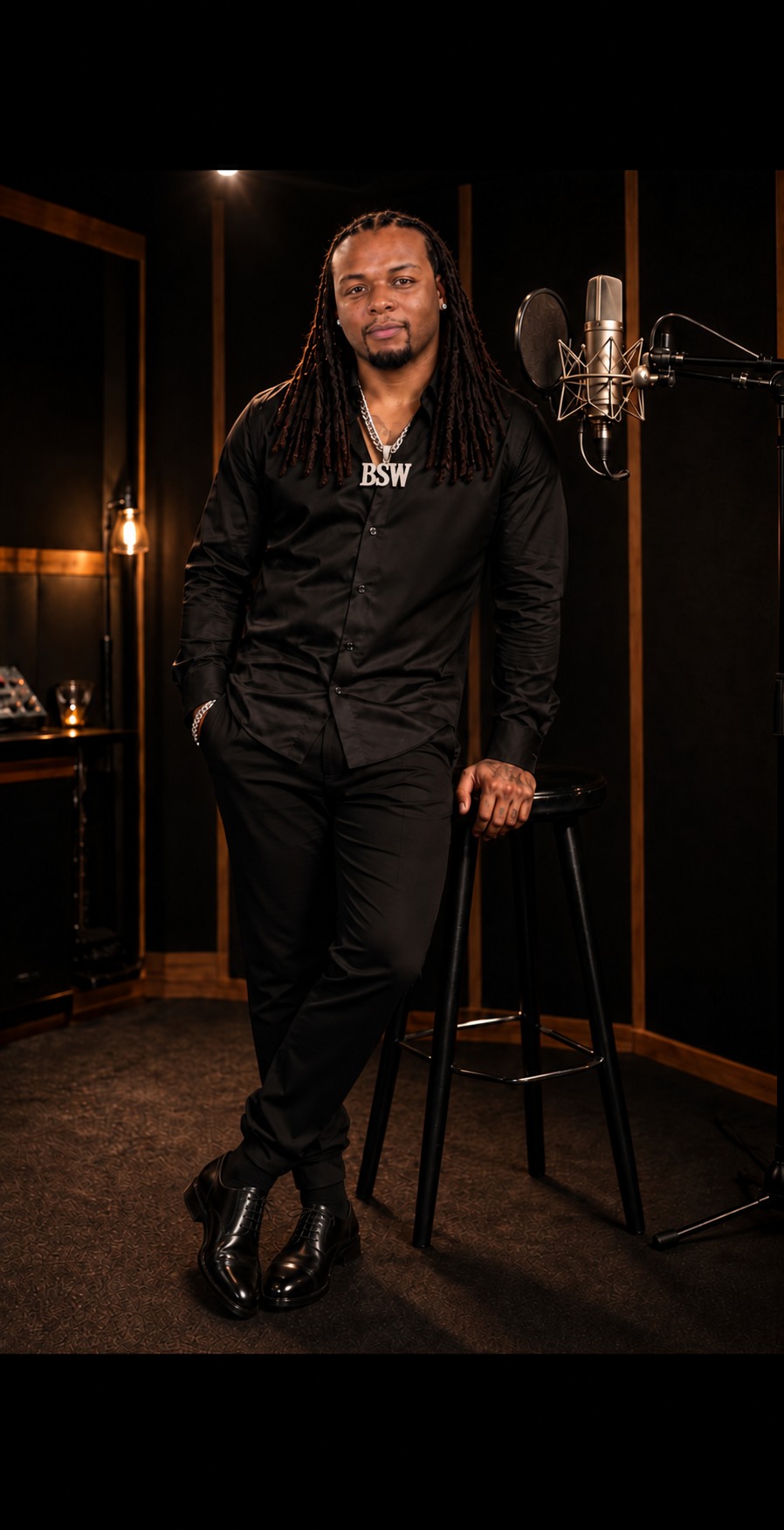 Coach Uncle Ricky beside a studio microphone, wearing a black dress shirt, trousers, polished black Oxford dress shoes, and his BSW chain